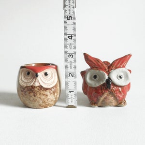 May include: Two ceramic owl figurines, one brown with red accents and the other red with white accents. Both owls have black eyes and are approximately 2 inches tall.