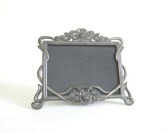 May include: A rectangular, pewter-colored picture frame with a floral design. The frame features roses at the top and bottom, with curved lines and swirls. The frame is designed to hold a photograph or artwork.