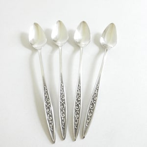 May include: Four silver spoons with elongated bowls and ornate, engraved handles. The spoons are arranged on a white surface, casting subtle shadows. The handles feature a detailed, scrolling floral design, adding a touch of elegance to the silverware.