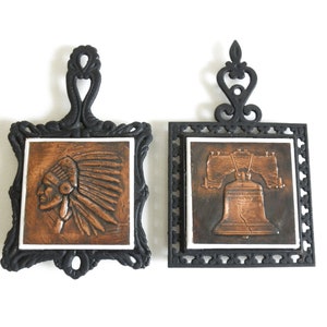 May include: Two black cast iron trivets with copper-colored ceramic tiles. One tile features a profile of a Native American wearing a headdress. The other tile features a copper-colored Liberty Bell.