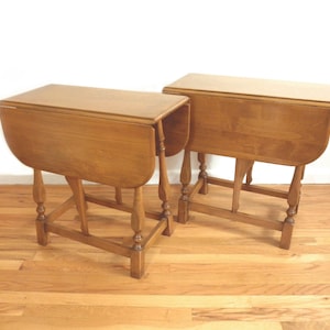 May include: Two wooden drop-leaf tables with a light brown finish. Each table has a rectangular top with rounded edges and turned legs. The tables are set on a hardwood floor.