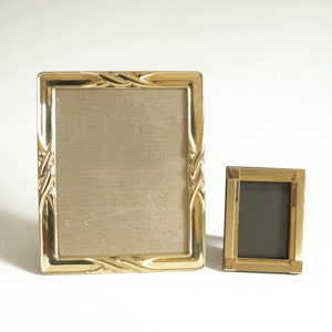 May include: Two gold picture frames, one larger than the other. The larger frame has a decorative, ornate design. Both frames have a gold finish.