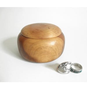 May include: A wooden jewelry box with a rounded shape and a lid. The box is made of light brown wood. Two rings are next to the box. One ring has a silver filigree design, and the other has a turquoise inlay.