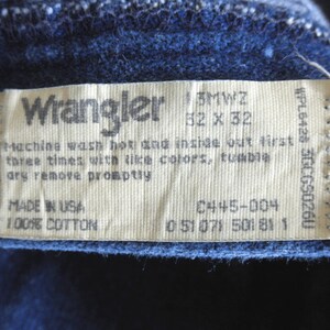 May include: A white Wrangler clothing care label with black text. The label includes the size 32 x 32, the style number 0445-004, and the fabric content 100% cotton. The label also includes washing instructions: machine wash hot and inside out first three times with like colours, tumble dry remove promptly. The label is made in the USA.