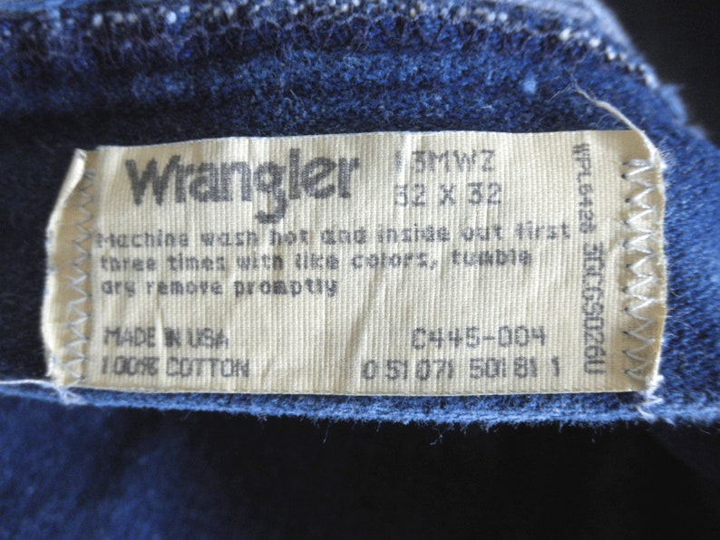 May include: A white Wrangler clothing care label with black text. The label includes the size 32 x 32, the style number 0445-004, and the fabric content 100% cotton. The label also includes washing instructions: machine wash hot and inside out first three times with like colours, tumble dry remove promptly. The label is made in the USA.