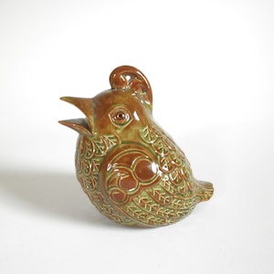 May include: A ceramic bird figurine with a green and brown glaze. The bird has a detailed design of leaves and a raised crest on its head.