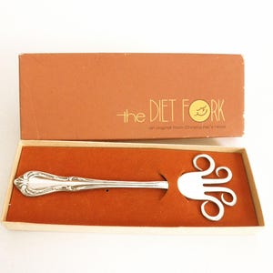 May include: A vintage silver-toned "Diet Fork" in its original box. The fork has a decorative handle and a unique, open-work design at the tines. The box is brown with the text "the DIET FORK".