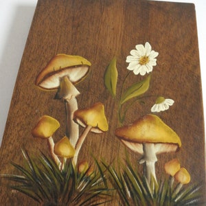 Puede incluir: Una placa de madera con un asa de cuerda presenta una escena pintada a mano de setas amarillas y flores blancas sobre un fondo marr&oacute;n. La placa est&aacute; firmada "Tracy".
