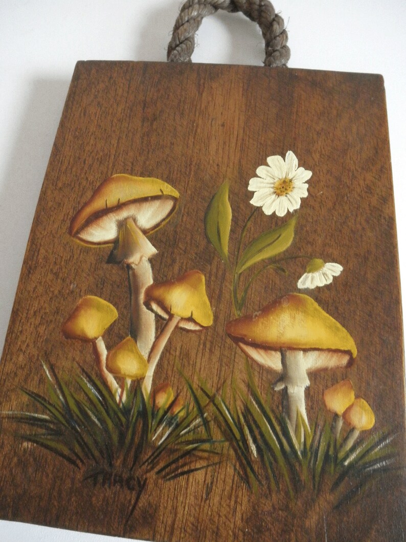 Puede incluir: Una placa de madera con un asa de cuerda presenta una escena pintada a mano de setas amarillas y flores blancas sobre un fondo marr&oacute;n. La placa est&aacute; firmada "Tracy".