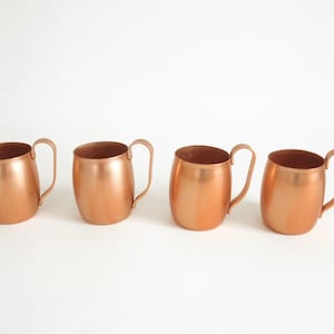 May include: Four copper mugs with handles. The mugs are all the same size and shape, and they are arranged in a row.