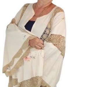 May include: A cream-colored dress with intricate lace and floral embroidery. The dress features wide sleeves and a flowing skirt, with a contrasting dark blue underlayer visible at the neckline. The dress is worn by a person wearing glasses.