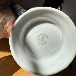 Puede incluir: Jarra de cer&aacute;mica blanca con asa y boquilla negras. La base de la jarra tiene un sello verde descolorido con texto y un dise&ntilde;o. La jarra est&aacute; sobre una superficie de madera.