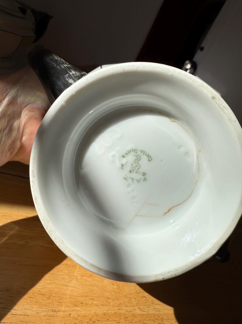 Puede incluir: Jarra de cer&aacute;mica blanca con asa y boquilla negras. La base de la jarra tiene un sello verde descolorido con texto y un dise&ntilde;o. La jarra est&aacute; sobre una superficie de madera.