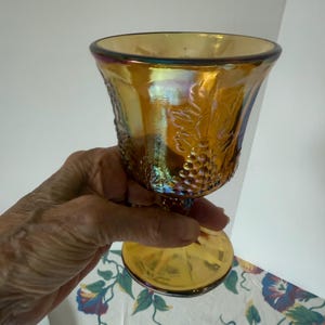 May include: An iridescent amber-colored goblet with a pedestal base. The glass features a grape vine design and a flared rim. The goblet is held up against a white background with a floral pattern.
