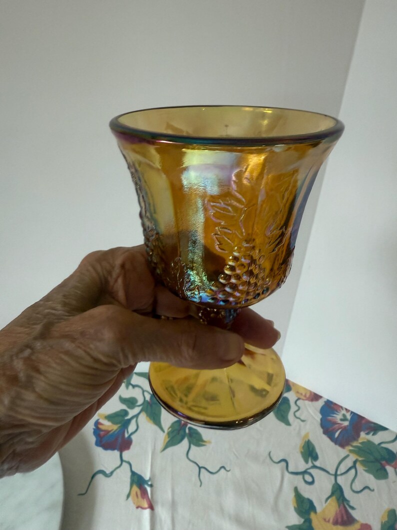 May include: An iridescent amber-colored goblet with a pedestal base. The glass features a grape vine design and a flared rim. The goblet is held up against a white background with a floral pattern.