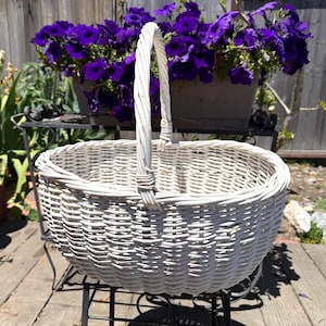 May include: An oval-shaped white wicker basket featuring a single tall arched handle attached to the center of the rim. The basket is constructed with a woven pattern of thin, flexible branches, creating a sturdy and open container design.
