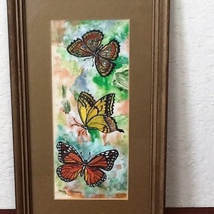 May include: A framed watercolor painting of three butterflies. The butterflies are in shades of orange, yellow, and brown. The background is a watercolor wash of green, blue, and orange.