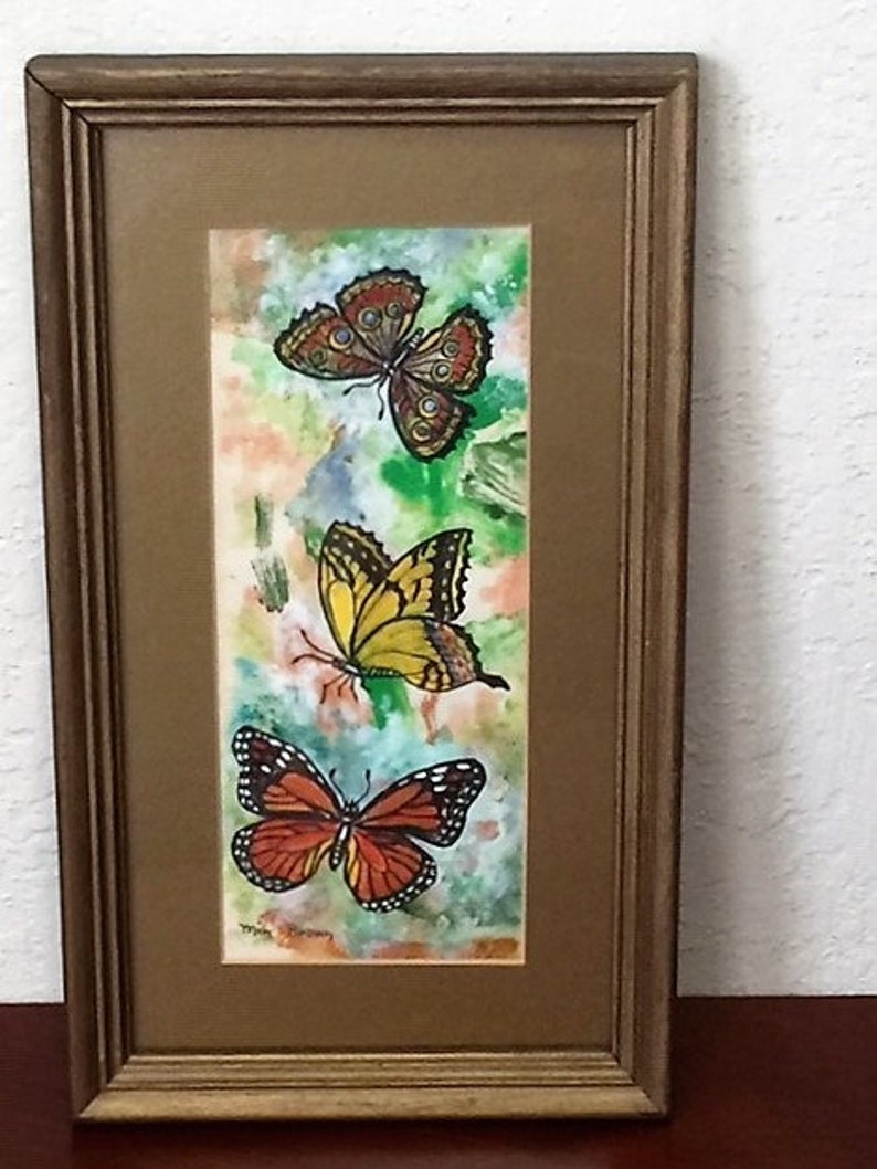 May include: A framed watercolor painting of three butterflies. The butterflies are in shades of orange, yellow, and brown. The background is a watercolor wash of green, blue, and orange.