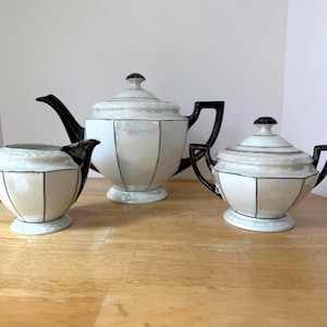 Può includere: Un servizio da tè in tre pezzi con finitura perlata. Il set include una teiera, una lattiera e una zuccheriera, tutti con dettagli e manici neri. Il design presenta linee nere verticali, creando un motivo geometrico su ogni pezzo.