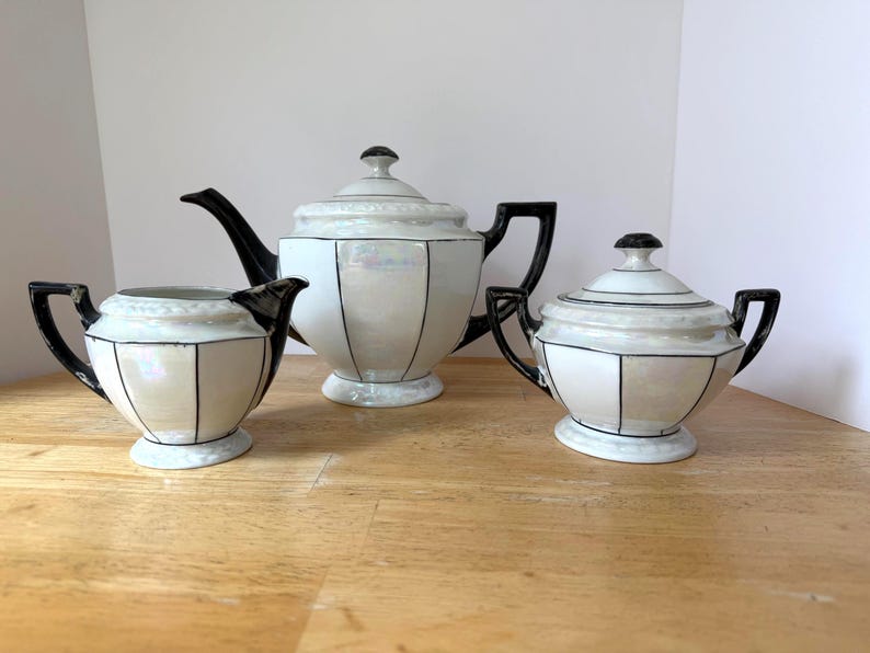 Puede incluir: Un juego de t&eacute; de tres piezas con acabado nacarado. El juego incluye una tetera, una crema y un azucarero, todos con detalles y asas negros. El dise&ntilde;o presenta l&iacute;neas negras verticales, creando un patr&oacute;n geom&eacute;trico en cada pieza.