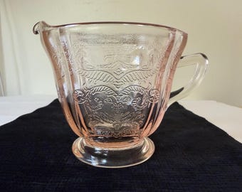 Puede incluir: Una cremera de vidrio rosa vintage con un diseño floral en relieve. La cremera tiene un asa y un pico, y se asienta sobre una base redonda. El vidrio es transparente, permitiendo el paso de la luz, y está sobre un fondo oscuro.