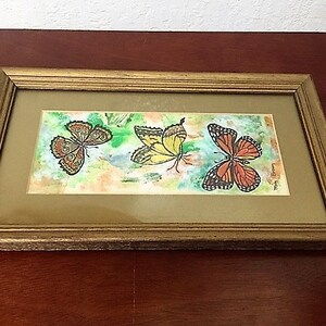 May include: A framed watercolor painting of three butterflies. The butterflies are yellow, orange, and brown with black markings. The background is a light wash of blue and green.