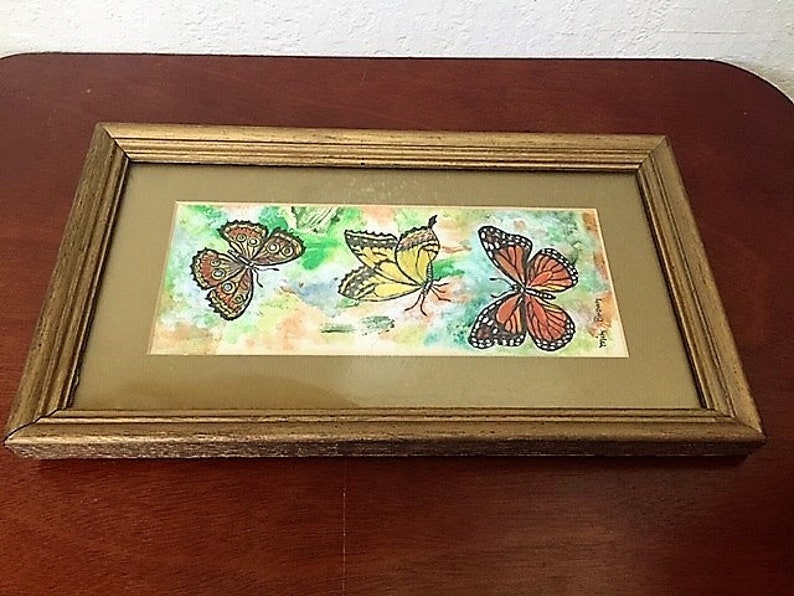 May include: A framed watercolor painting of three butterflies. The butterflies are yellow, orange, and brown with black markings. The background is a light wash of blue and green.
