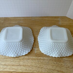 May include: Two white milk glass bowls with a square bottom and a diamond pattern on the sides. The bowls are empty and sitting on a wooden surface.