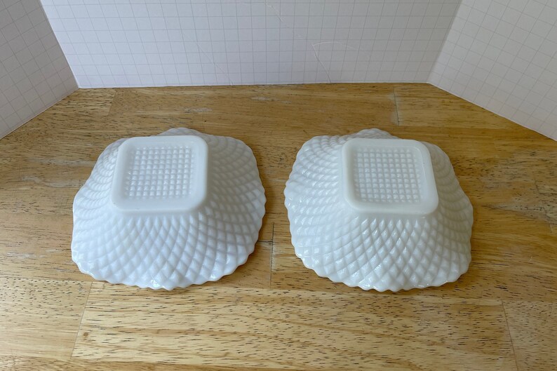 May include: Two white milk glass bowls with a square bottom and a diamond pattern on the sides. The bowls are empty and sitting on a wooden surface.