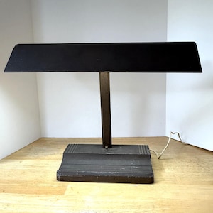 May include: A vintage black desk lamp with a rectangular shade and a dark metal base. The lamp has a vertical support and a horizontal shade, and a power cord extends from the base. The lamp is designed for desk use.
