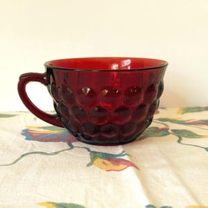 May include: A ruby red glass teacup with a handle. The cup has a textured, bubble-like pattern on the exterior. The cup is sitting on a floral patterned tablecloth.