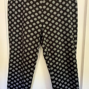 May include: Black pants featuring a repeating white circular pattern with a central dot and a radiating sunburst design. The pants have a straight-leg cut, a high waistband, and are displayed on a plastic hanger against a white door.