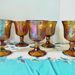 May include: Six iridescent amber-colored goblets with a grape design. The goblets have a flared bowl, a short stem, and a round base. The glass reflects a rainbow of colors. The goblets are arranged on a floral patterned tablecloth.
