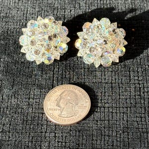 Puede incluir: Dos pendientes claros y brillantes en forma de flor con cristales iridiscentes. Los pendientes est&aacute;n junto a una moneda de veinticinco centavos de d&oacute;lar estadounidense. Los pendientes est&aacute;n sobre una superficie negra texturizada.