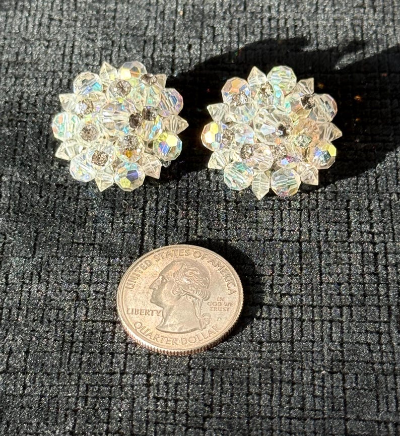 Puede incluir: Dos pendientes claros y brillantes en forma de flor con cristales iridiscentes. Los pendientes est&aacute;n junto a una moneda de veinticinco centavos de d&oacute;lar estadounidense. Los pendientes est&aacute;n sobre una superficie negra texturizada.