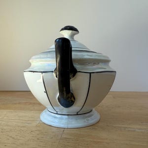 Puede incluir: Una tetera de cer&aacute;mica blanca con asa y detalles en negro. La tetera tiene un cuerpo redondeado, una tapa con un pomo negro y est&aacute; decorada con l&iacute;neas negras. La tetera se asienta sobre una base blanca.