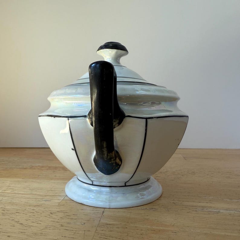 Puede incluir: Una tetera de cer&aacute;mica blanca con asa y detalles en negro. La tetera tiene un cuerpo redondeado, una tapa con un pomo negro y est&aacute; decorada con l&iacute;neas negras. La tetera se asienta sobre una base blanca.