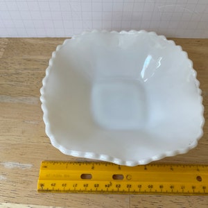 May include: A white, square-shaped, milk glass dish with a scalloped edge. The dish is approximately 5 inches wide.