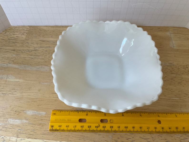 May include: A white, square-shaped, milk glass dish with a scalloped edge. The dish is approximately 5 inches wide.