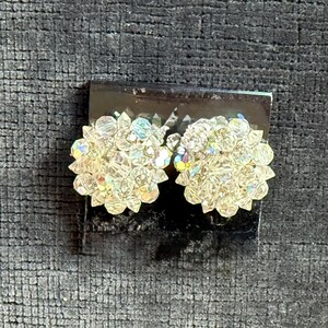 Puede incluir: Un par de pendientes de clip de cristal transparente y brillante. Cada pendiente es un grupo de peque&ntilde;os cristales facetados dispuestos en un dise&ntilde;o floral. Los cristales reflejan la luz, creando un efecto arco&iacute;ris. Los pendientes est&aacute;n sobre una base oscura.