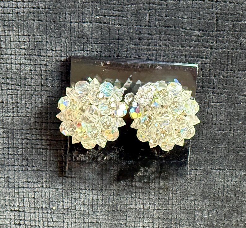 Puede incluir: Un par de pendientes de clip de cristal transparente y brillante. Cada pendiente es un grupo de peque&ntilde;os cristales facetados dispuestos en un dise&ntilde;o floral. Los cristales reflejan la luz, creando un efecto arco&iacute;ris. Los pendientes est&aacute;n sobre una base oscura.