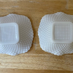 May include: Two white milk glass bowls with a square shape and a raised, textured design. The bowls are empty and sitting on a wooden surface.