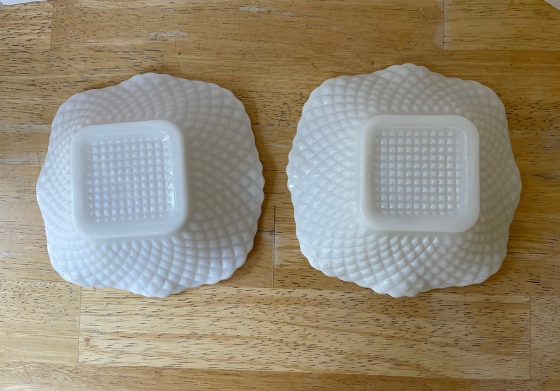 May include: Two white milk glass bowls with a square shape and a raised, textured design. The bowls are empty and sitting on a wooden surface.