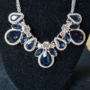 May include: A silver-toned statement necklace with large, dark blue teardrop and oval-shaped gemstones. The gemstones are surrounded by smaller, clear crystals, creating a sparkling effect. The necklace is displayed on a black velvet surface.