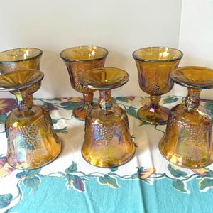 May include: Six iridescent amber-colored goblets with a bell-shaped base and a round top. The goblets have a textured surface with a grape design. The stem is short and has a decorative design.