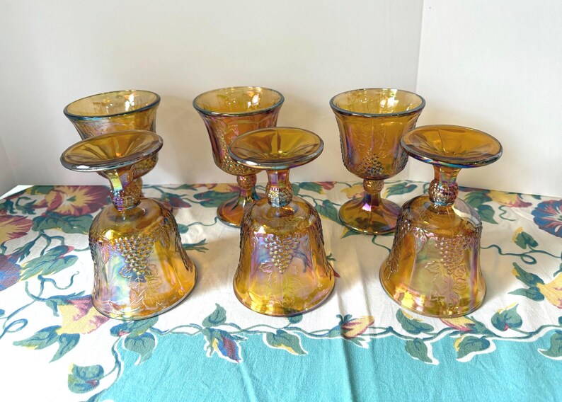 May include: Six iridescent amber-colored goblets with a bell-shaped base and a round top. The goblets have a textured surface with a grape design. The stem is short and has a decorative design.