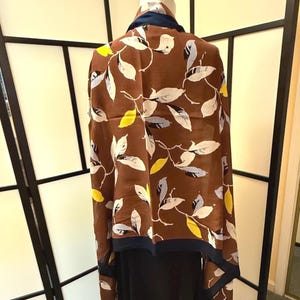 May include: A brown scarf featuring a repeating pattern of white, light blue, and yellow leaves. The scarf has a solid dark navy blue border along the edges and is draped over a mannequin, showing a smooth, lightweight fabric texture throughout.