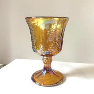 May include: An iridescent amber-colored goblet with a detailed grape vine design. The glass has a flared rim, a short stem, and a round base. The goblet's surface reflects a rainbow of colors, adding to its vintage charm.