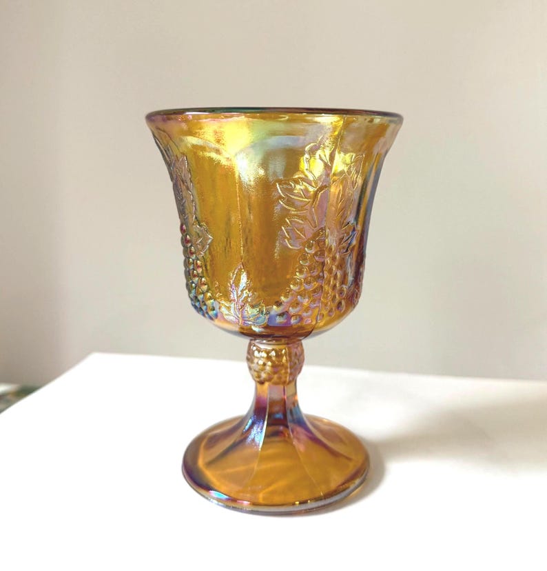 May include: An iridescent amber-colored goblet with a detailed grape vine design. The glass has a flared rim, a short stem, and a round base. The goblet's surface reflects a rainbow of colors, adding to its vintage charm.