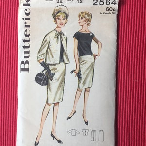 Pu&ograve; includere: Una busta per cartamodello Butterick, numero 2564, taglia 12, busto 32. L'illustrazione mostra due figure che indossano un completo gonna e giacca abbinato, con un top scuro, guanti neri e una borsa. Include disegni al tratto dei capi.